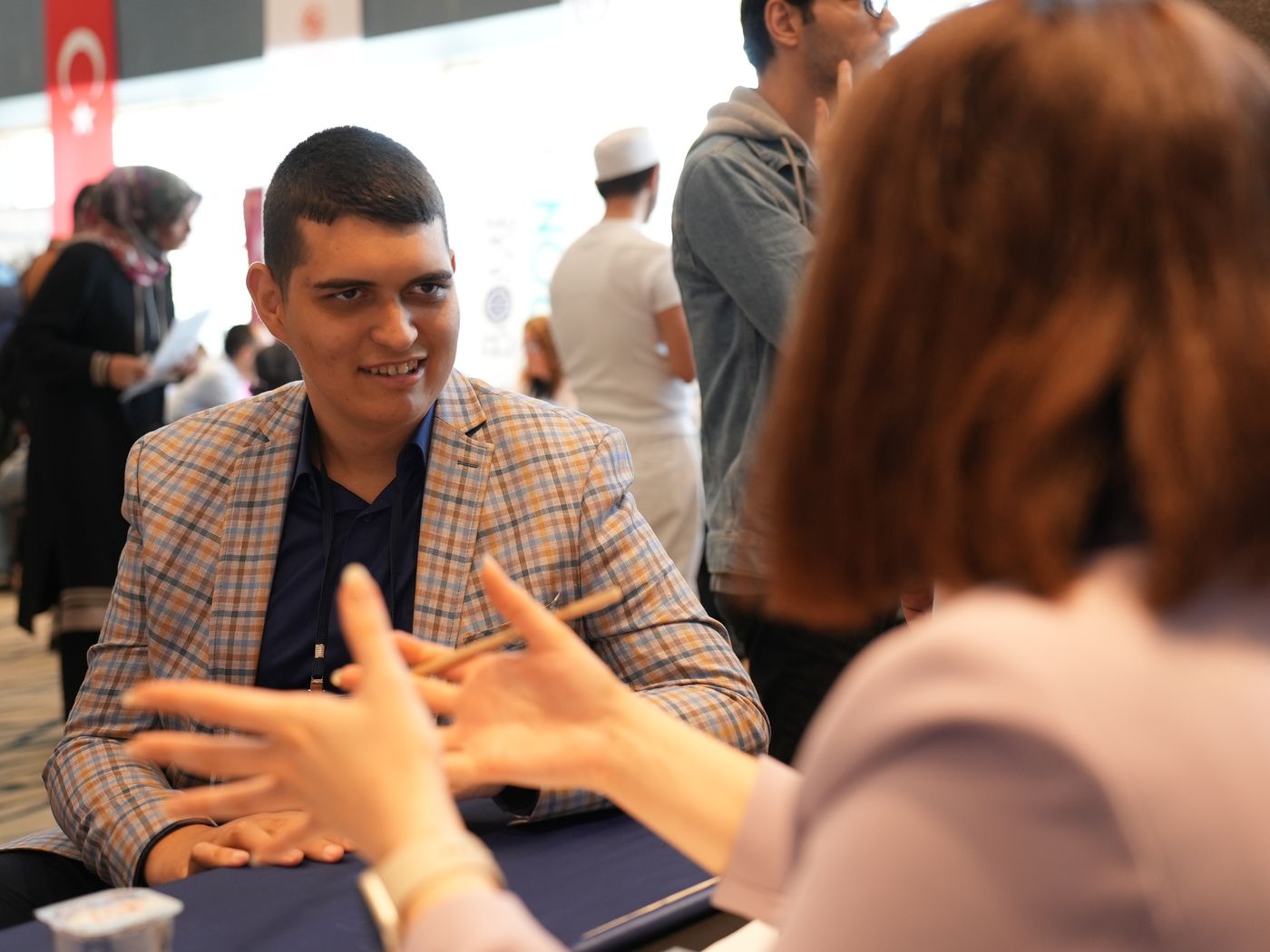 A young man in a checked blazer sits at a table in conversation with a woman, both gesturing as they talk at a disability employment event.