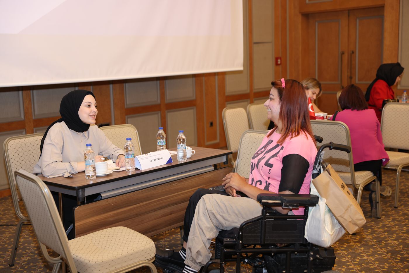Two women talk across a table at a job fair. One uses a powered wheelchair; both are smiling mid-conversation.