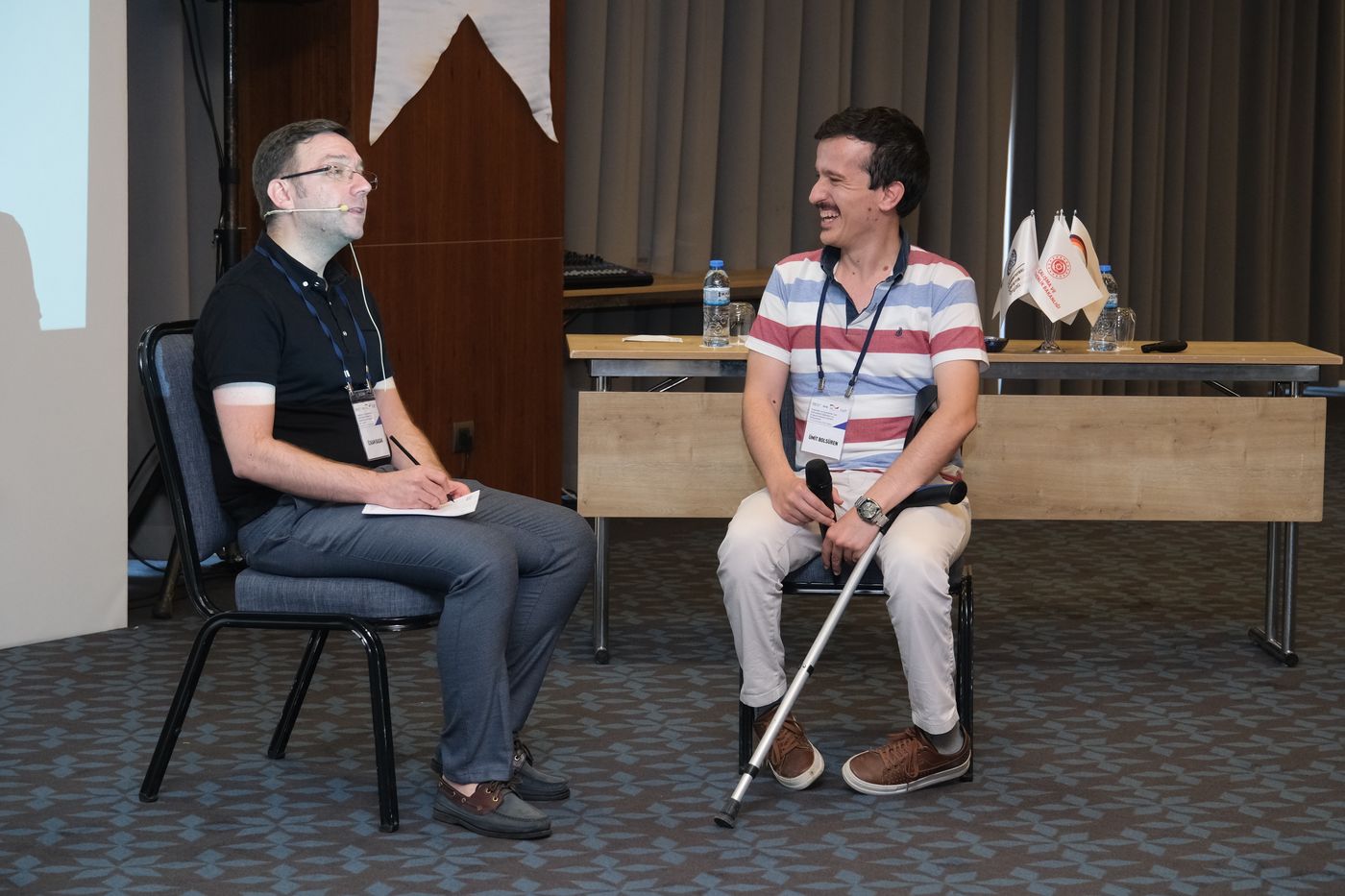 Two men sit facing each other on a stage. One holds a microphone and a walking crutch rests against his chair; the other takes notes.